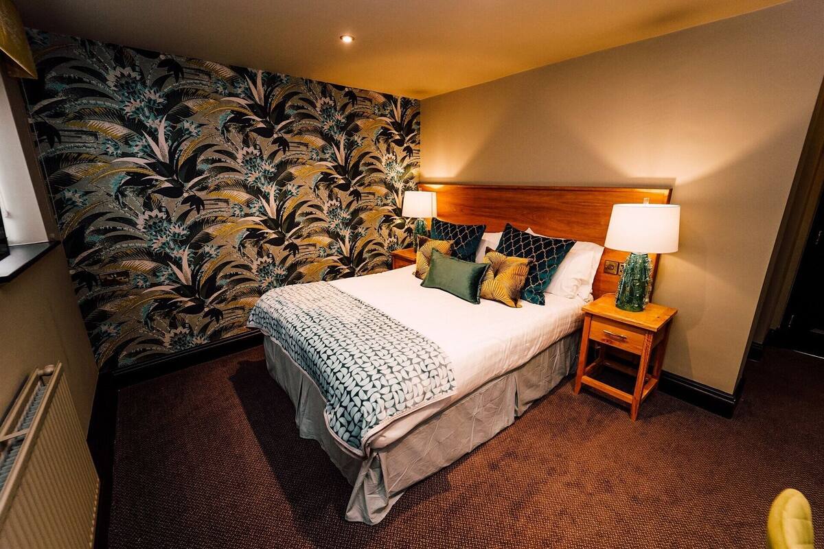 A hotel room with a bed, two bedside tables with lamps, a large floral wallpaper, and a radiator.