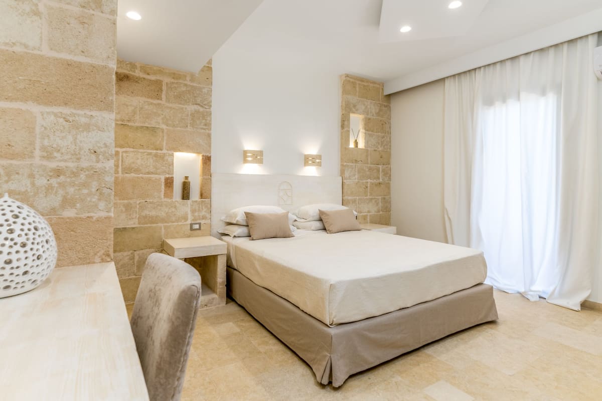 A modern hotel room with a stone accent wall, a large bed with beige bedding, a bedside table with a lamp, and a large window with curtains.