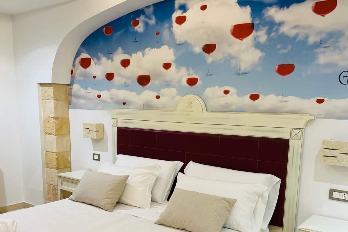 A hotel room with a large bed, a headboard with a decorative design, and a mural of clouds and hot air balloons on the wall.