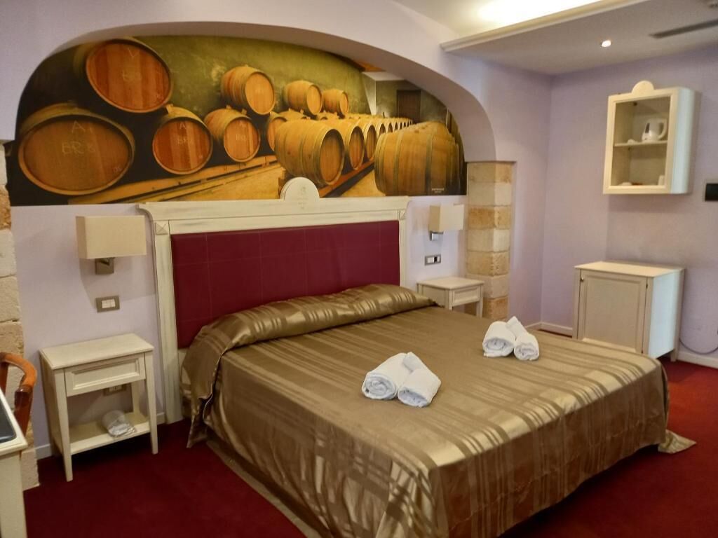Habitación de hotel con una cama grande, cabecera roja y un mural con barricas de vino en la pared. Hay dos mesitas de noche, una mesilla auxiliar y un armario pequeño.