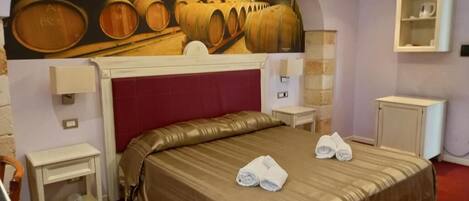 Habitación de hotel con una cama grande, cabecera roja y un mural con barricas de vino en la pared. Hay dos mesitas de noche, una mesilla auxiliar y un armario pequeño.