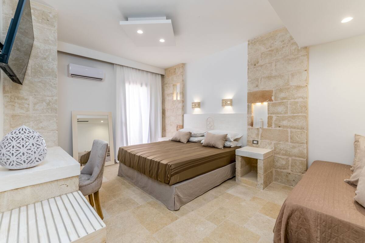 A modern bedroom with a large bed, a desk, and a chair. There is a stone wall and a mounted TV.