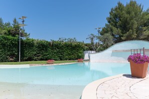Ein Schwimmbecken unter blauem Himmel, umgeben von viel Grün und einer weißen Mauer.