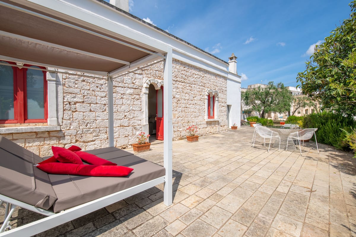 A stone building with a covered patio area, a lounge chair with red cushions, and a table with chairs.
