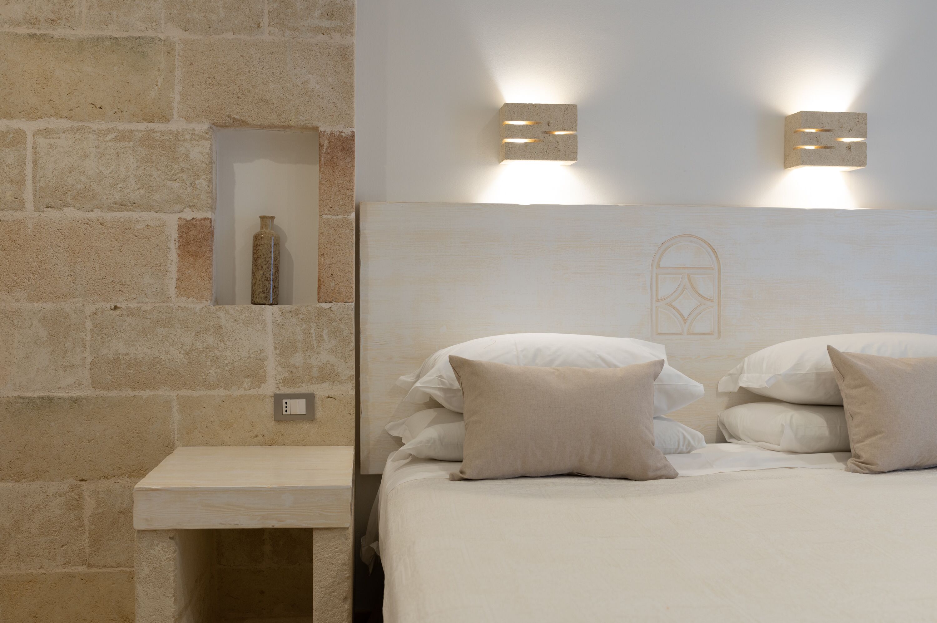 A bedroom with a stone wall, a bedside table with a bottle, and two wall-mounted light fixtures.