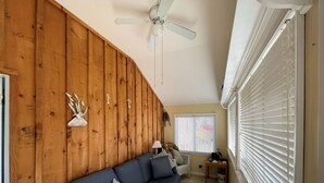 A living room with a wooden wall, a blue sofa, a ceiling fan, and a window with blinds.