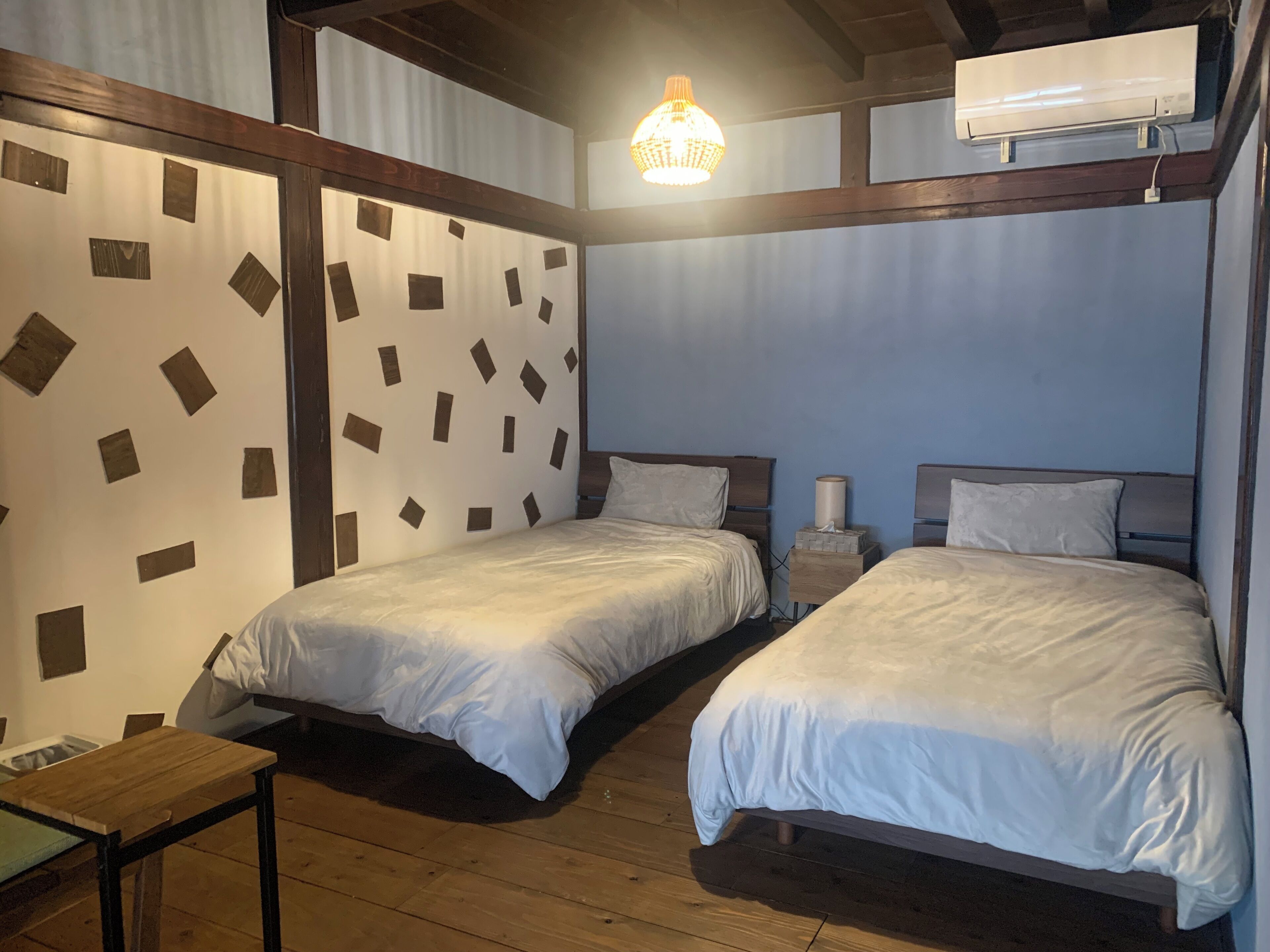 Quarto de hotel com duas camas, piso de madeira, uma mesinha e uma parede com padrões geométricos.