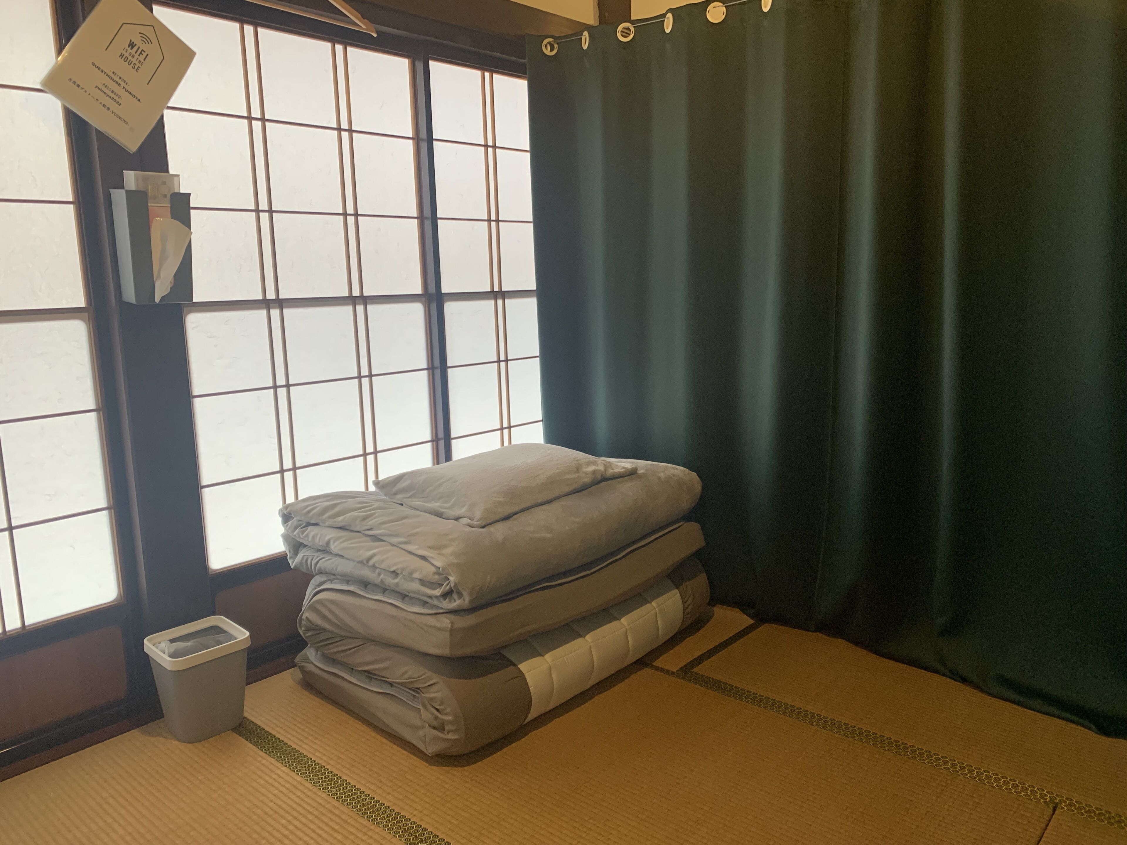 A room with tatami flooring, a bed with a grey mattress, a trash bin, and a window with a curtain.