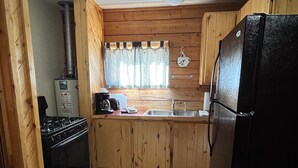 A compact kitchen with wooden cabinets, a black refrigerator, a stove, a coffee maker, and a window with checkered curtains. - One Bedroom Cabin with Ample Parking and Space from Neighboring Cabins (Almont)