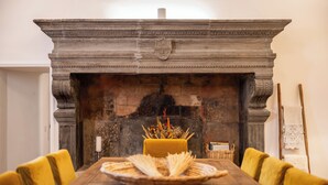 Una mesa de comedor con sillas amarillas, una chimenea con repisa decorativa y un jarrón con flores secas.