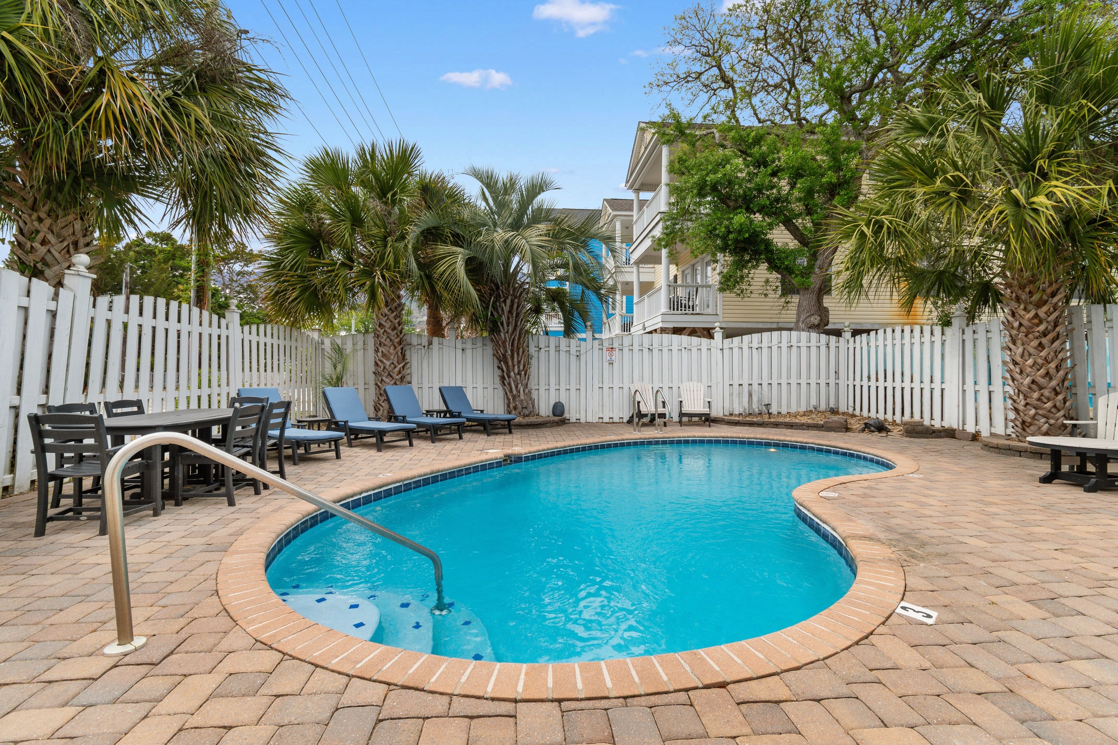 A swimming pool with a white fence, lounge chairs, and palm trees.
