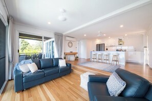 A modern living room with a sofa, a dining area with a kitchen in the background, and a balcony with curtains. - Airlie On Airlie Premier (Airlie Beach)