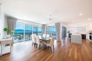 A spacious living area with a dining table and chairs, a kitchen, and large windows offering a view of the sea. - Airlie On Airlie Premier (Airlie Beach)