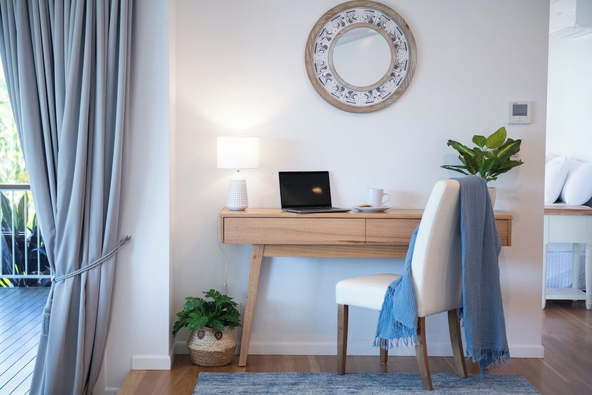 Quarto com escrivaninha, cadeira, laptop, luminária, planta em vaso e espelho decorativo.