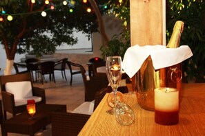 A table with a bottle of champagne, a glass, and a candle, set outdoors with string lights and chairs. - Nancy (Hersonissos)