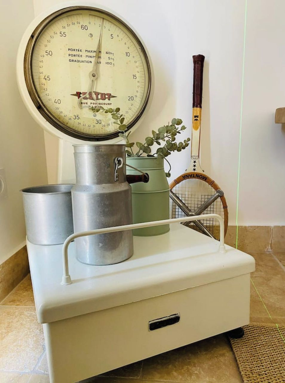 Une balance ancienne, un récipient en métal, un arrosoir vert et une raquette de tennis, le tout posé sur un meuble blanc.