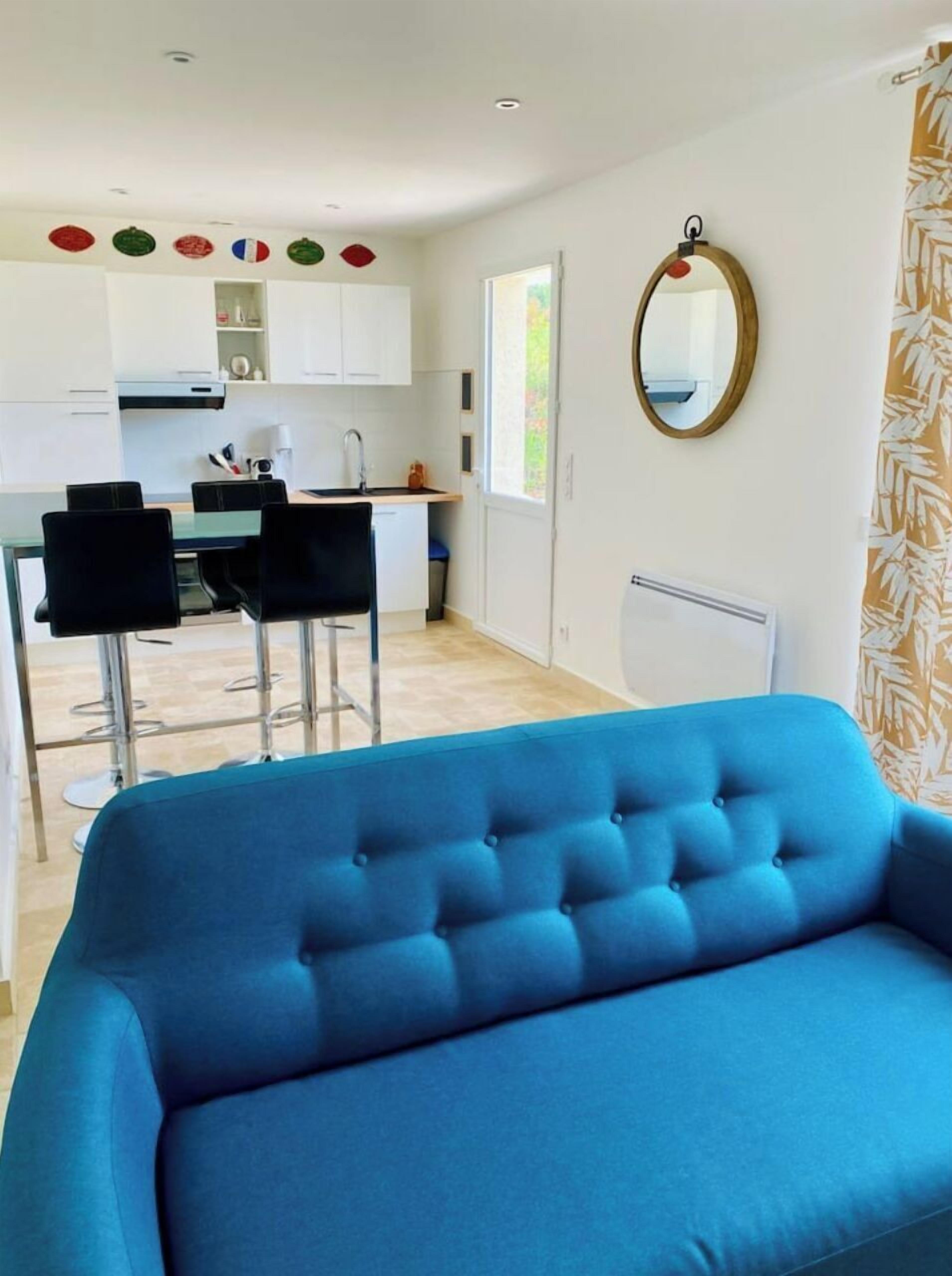 Un salon moderne avec un canapé bleu, un coin cuisine doté de meubles blancs et un espace repas avec des chaises noires.