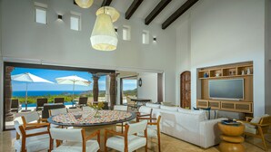 A spacious living area with a large round table, white sofas, and wooden chairs. A flat-screen TV is mounted on the wall, and there are multiple windows offering a view of the ocean.