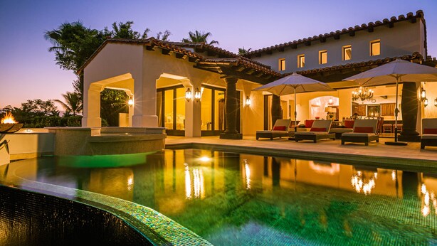 A pool area with a clear view of the pool, lounge chairs, and a building with a tiled roof.