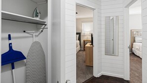 Ein Flur mit Einbauschrank, der Regale und abgehängte Kleidung beherbergt, einer Tür zu einem Schlafzimmer mit Bett und Schreibtisch sowie einem Blick in einen weiteren Raum mit Fenster und Lampe.