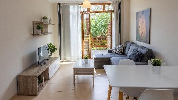Ein modernes Wohnzimmer mit einer Couch, einem Esstisch und einem Fernseher. Ein großes Fenster mit Vorhängen bietet Blick auf eine grüne Umgebung.