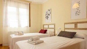Ein Hotelzimmer mit zwei Betten, jeweils mit einem hölzernen Kopfteil, weißer Bettwäsche und einem roten Kissen auf einem Bett. Auf den Betten liegen Handtücher, und es gibt ein Fenster mit durchsichtigen Vorhängen.