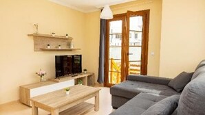Ein Wohnzimmer mit einer Couch, einem Holztisch, einem Fernseher auf einem Schrank und einem Fenster mit Vorhängen.