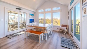A spacious dining area with a wooden table and white chairs, large windows with starfish decorations, and a view of the outside. - The BARNacle - Corolla, Ocean Sands, 5 Min. Walk to Beach, Pool, Hot Tub, Pets OK, Flex Stays (Corolla)