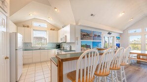 A spacious kitchen with white cabinets, a central island with bar stools, and artwork on the walls. - The BARNacle - Corolla, Ocean Sands, 5 Min. Walk to Beach, Pool, Hot Tub, Pets OK, Flex Stays (Corolla)