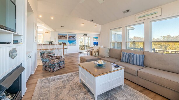 A spacious living room with a sofa, coffee table, and a fireplace. There is a dining area with chairs and a table, and a kitchen in the background. - The BARNacle - Corolla, Ocean Sands, 5 Min. Walk to Beach, Pool, Hot Tub, Pets OK, Flex Stays (Corolla)