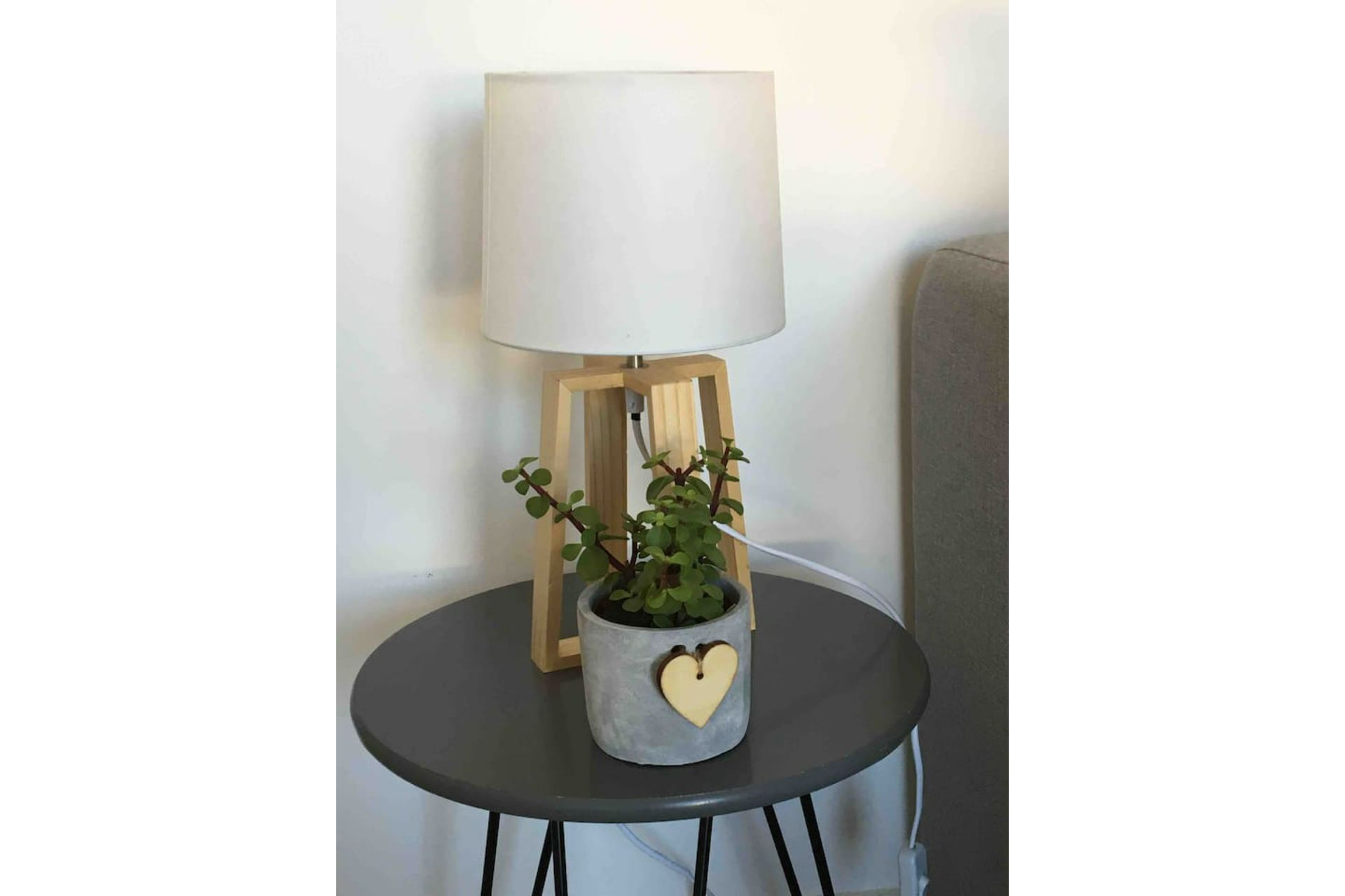 A round table with a grey pot containing a plant and a heart-shaped object. A lamp with a white shade is on a wooden stand behind the table.