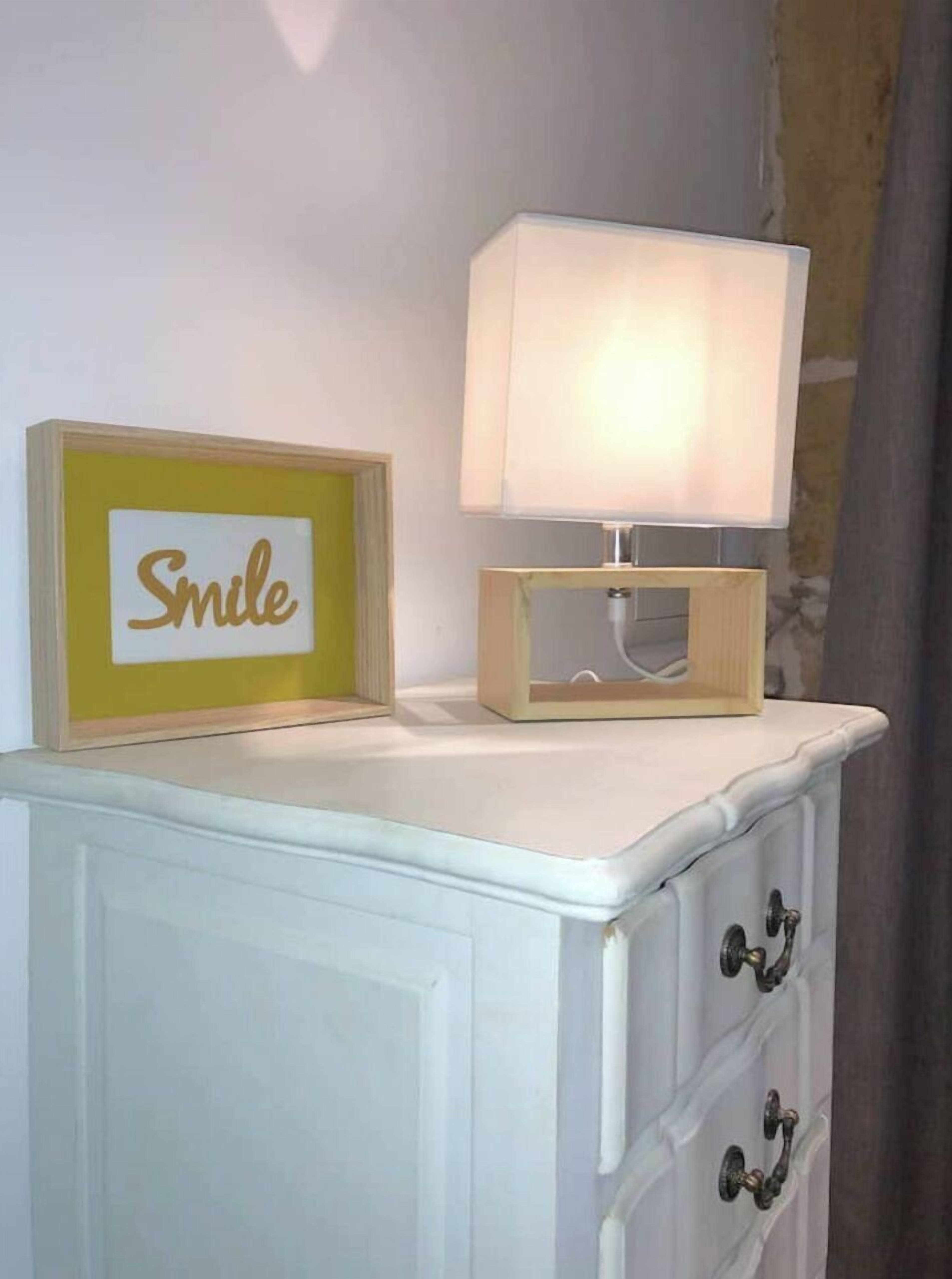 Une commode blanche sur laquelle est posé un cadre avec l’inscription « Souriez » et une lampe de table.