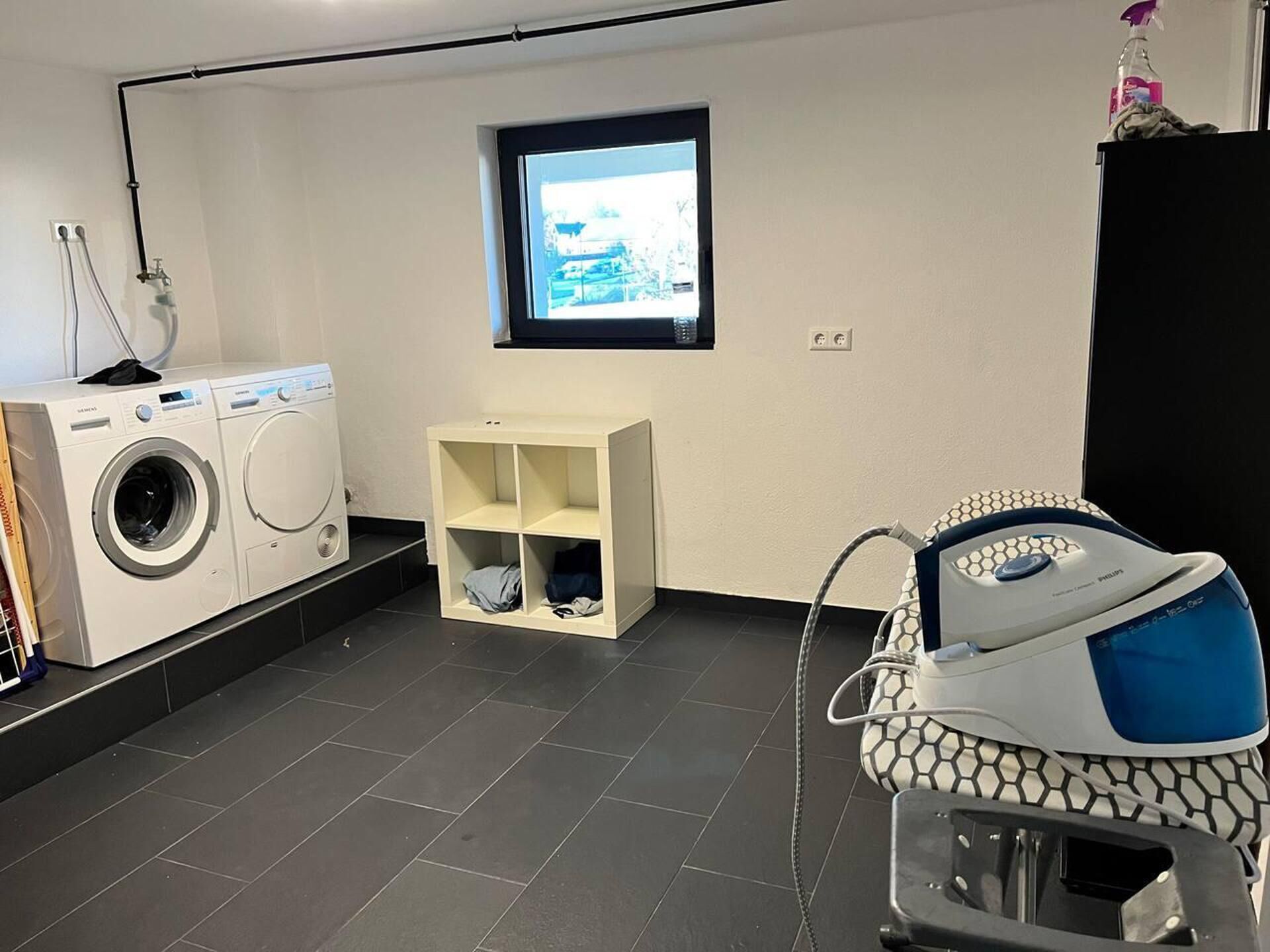 Een wasruimte met een wasmachine, een droogrek, een plank met opgevouwen kleding en een stofzuiger.