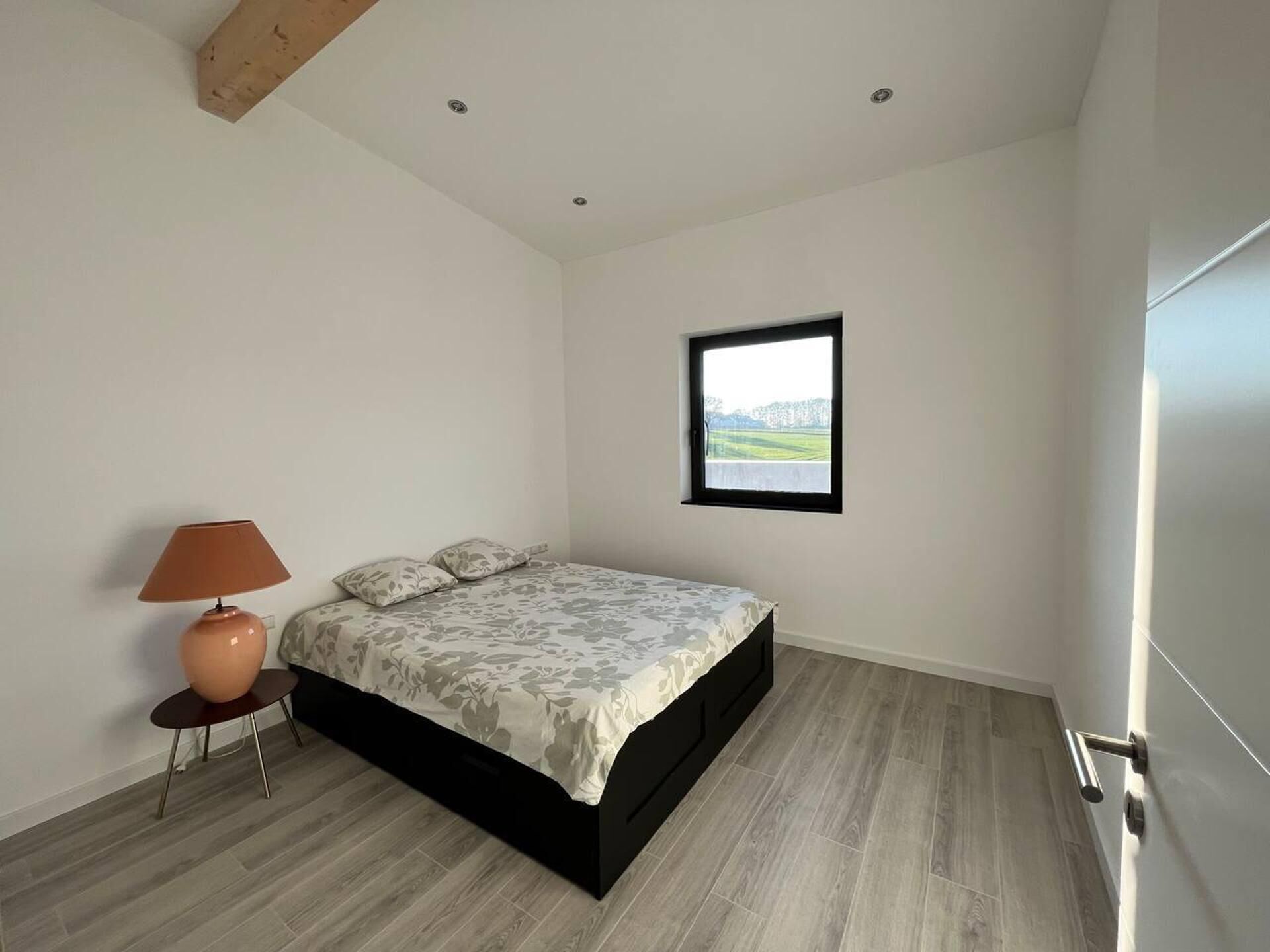 A bedroom with a bed, a nightstand, a lamp, a window with a view, and a door.