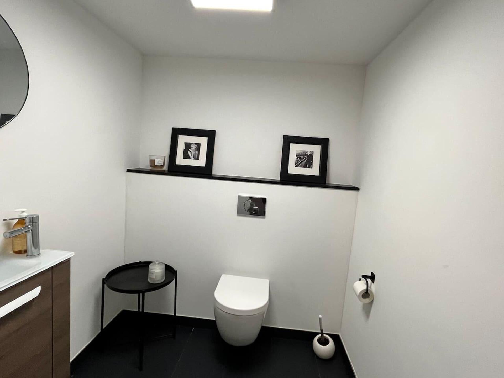 A small, modern bathroom with a white toilet, a small round table with a bottle, a shelf with two framed pictures, and a toilet brush.