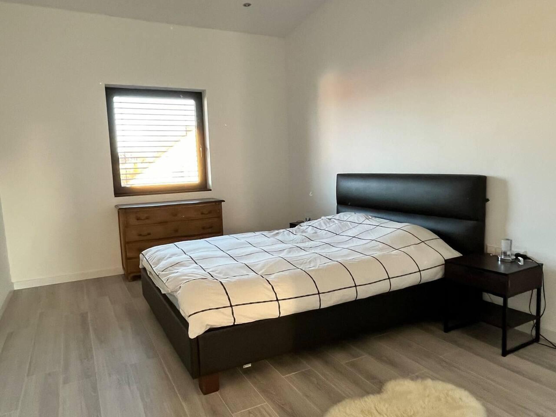 Une chambre à coucher comprenant un lit, une table de chevet, une commode et une fenêtre avec des stores.