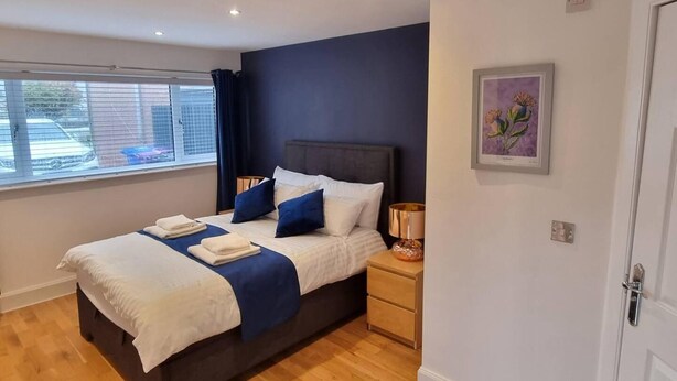 A modern bedroom with a large bed, two bedside tables, a window with blinds, and a door. - Laighdykes Guest Cottage 2 Bedroom & Gym Saltcoats (Saltcoats)