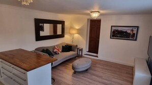 A modern living room with a sofa, ottoman, and a small table with a lamp. A large mirror and a framed picture are on the walls. - Laighdykes Guest Cottage 2 Bedroom & Gym Saltcoats (Saltcoats)