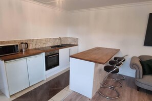 A compact kitchen with white cabinets, a wooden countertop, a built-in oven, a microwave, and a coffee maker. There is a small bar area with stools and a sink.