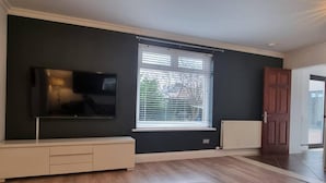 A living room with a flat-screen TV mounted on the wall, a window with blinds, a radiator, and a wooden floor. - Laighdykes Guest Cottage 2 Bedroom & Gym Saltcoats (Saltcoats)