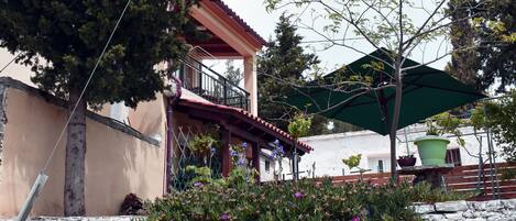 A stone wall with a bush of purple flowers, a building with a balcony, and a green umbrella.