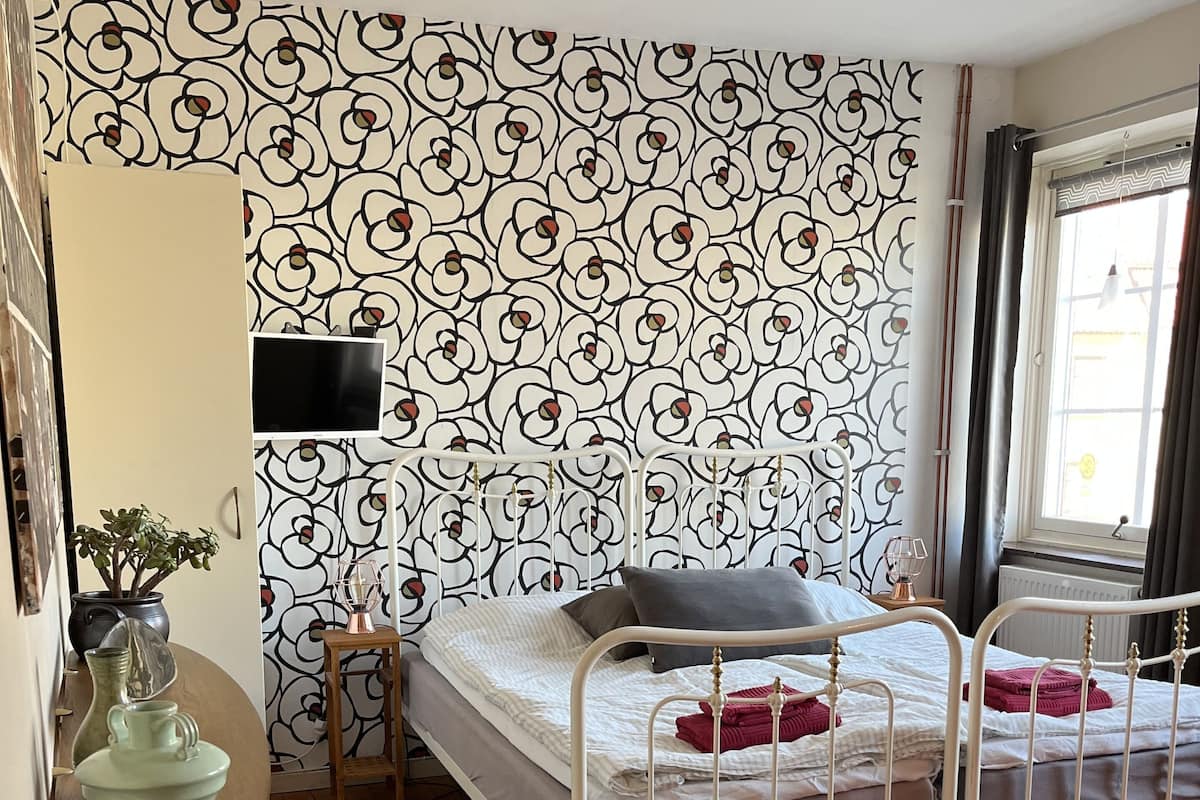 A bedroom with a patterned wallpaper, a bed with white metal frame, a red rug, and a small table with a plant.