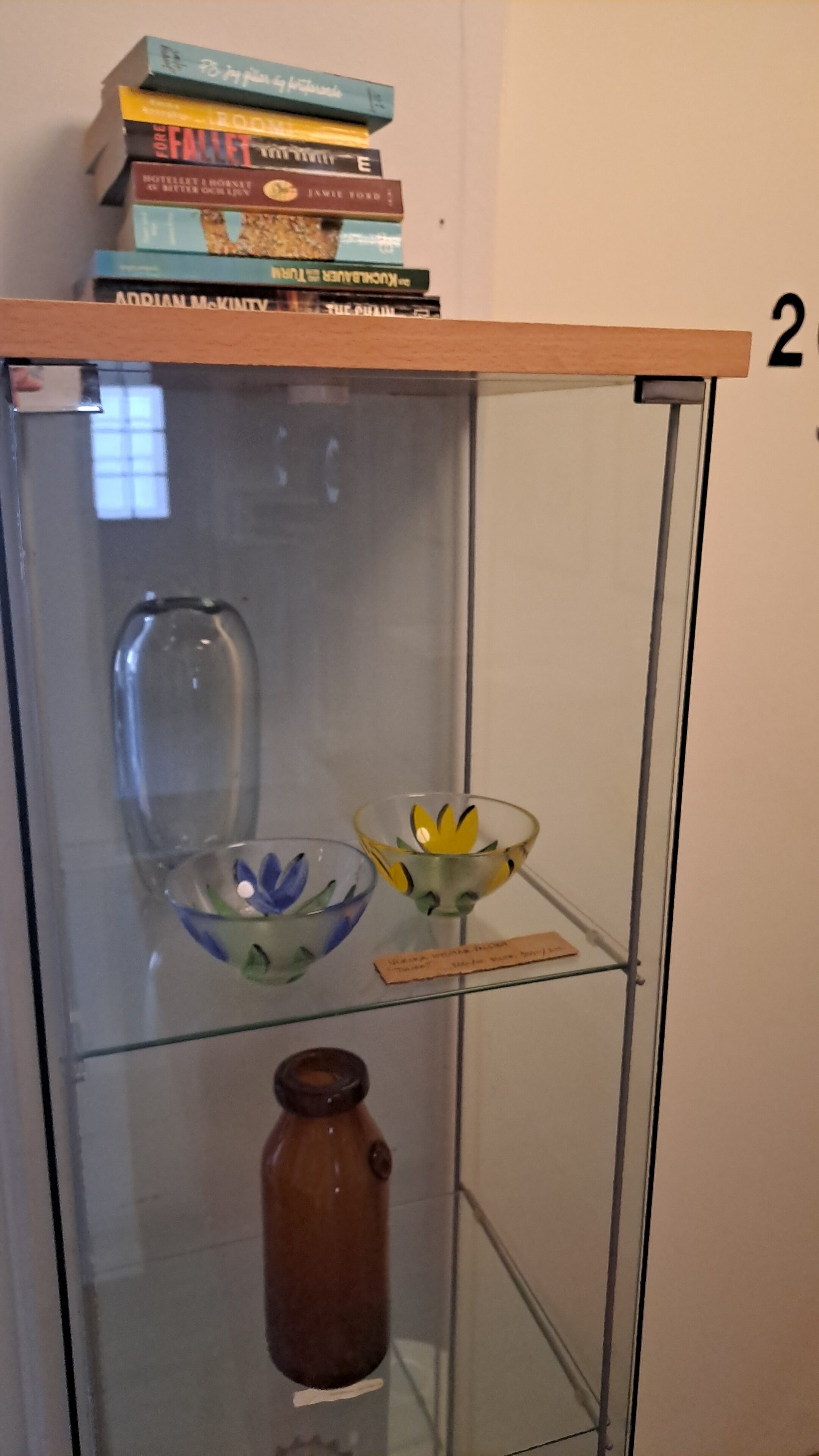 A glass display cabinet with a wooden shelf on top holding books. Inside, there are two glass bowls with flowers and a small brown vase.