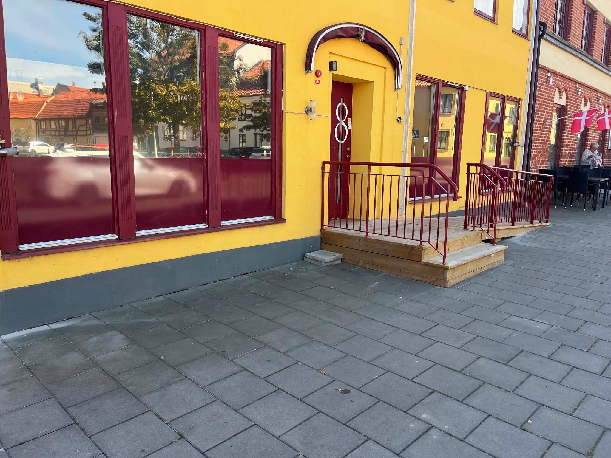 A yellow building with a red door and windows, a small porch with a red railing, and a paved sidewalk.
