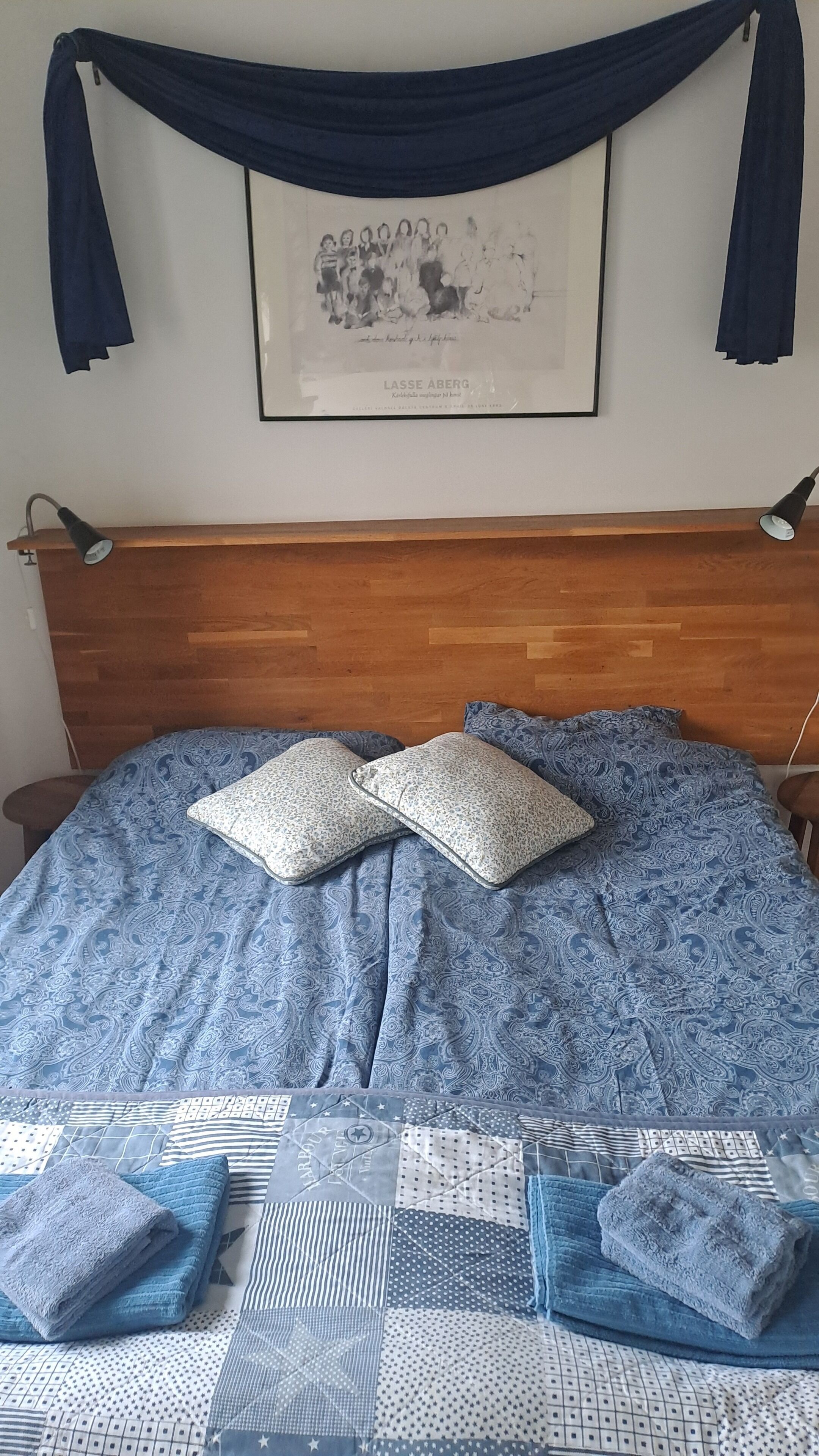 En säng med ett blått, mönstrat överkast, två kuddar och en inramad tavla ovanför.