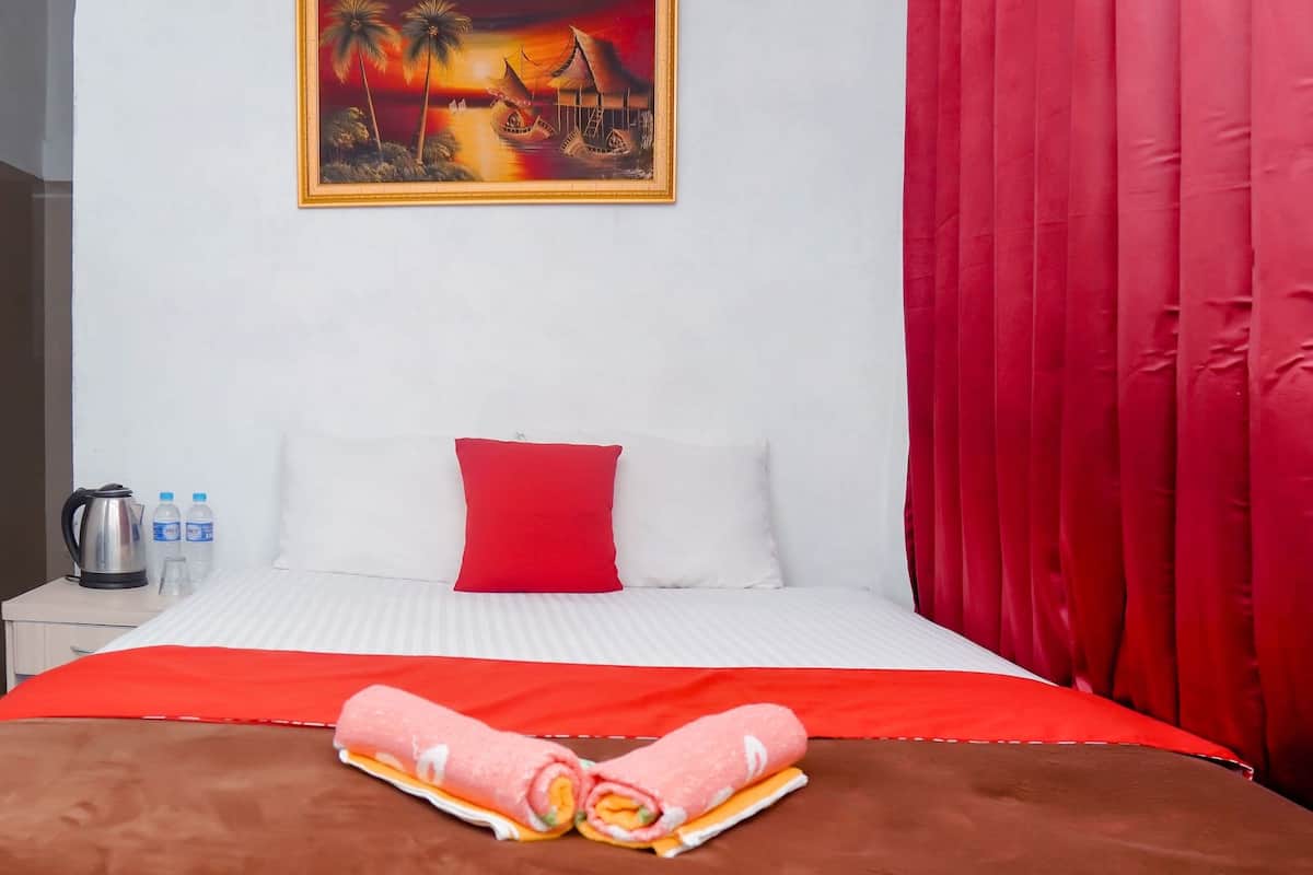 A hotel room with a bed, a red curtain, a painting of a sunset scene, and a small table with a kettle and water bottles.