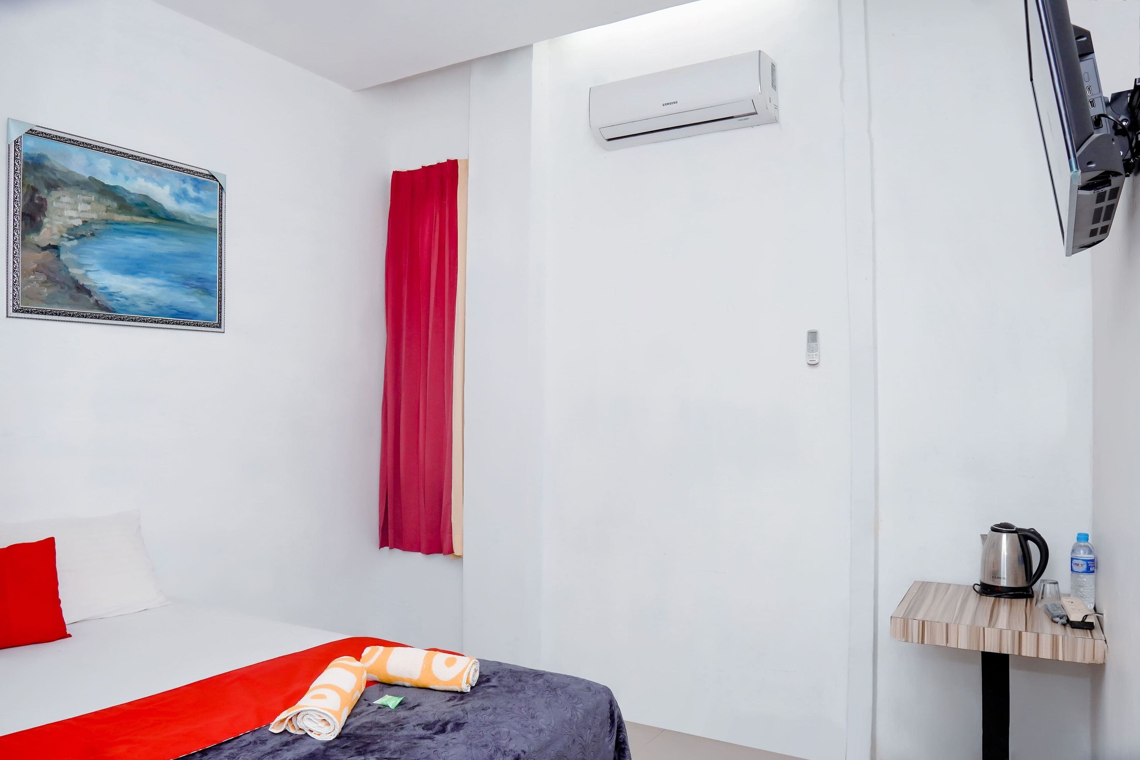 Une chambre d’hôtel avec un lit, un rideau rouge, un appareil de climatisation, une télévision et une petite table sur laquelle se trouvent une bouilloire et une bouteille d’eau.
