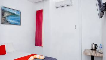 Une chambre d’hôtel avec un lit, un rideau rouge, un appareil de climatisation, une télévision et une petite table sur laquelle se trouvent une bouilloire et une bouteille d’eau.