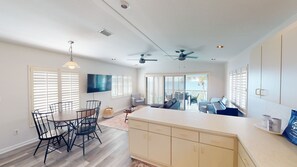Un grand espace de vie comprenant une table à manger, des chaises, un canapé et un téléviseur. On y trouve également des ventilateurs de plafond et une grande fenêtre équipée de stores.