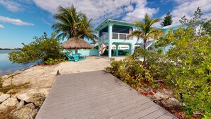 A beachside property with a wooden deck leading to a turquoise building, a thatched umbrella, and a flag. - Sea La Vie - Your Oceanfront Escape - 3BD/3Bath - Cudjoe Key (Cudjoe Key)